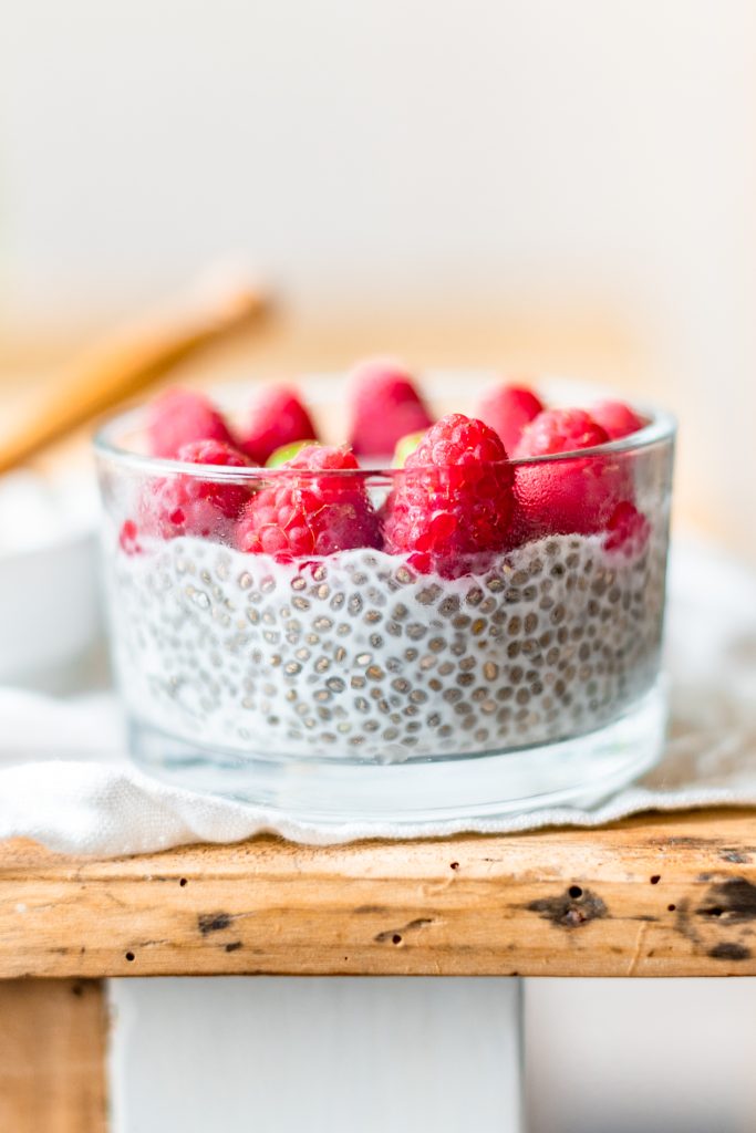 Astuces pour manger des graines au quotidien. Un bol en verre contenant du pudding de graines de chia au lait végétal, surmonté de framboises fraîches bien rouges. Le bol est posé sur une table en bois rustique, avec un linge blanc délicatement placé en dessous. L’arrière-plan est flou, mettant en valeur le dessert.