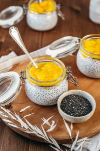 Astuces pour manger des graines au quotidien. Un dessert sain et appétissant composé de pudding de graines de chia au lait végétal, garni d'une purée de mangue. Le tout est présenté dans des petits bocaux en verre avec des couvercles métalliques, posés sur une planche en bois. Une cuillère est plongée dans l'un des bocaux, et un petit bol rempli de graines de chia est disposé à côté.
