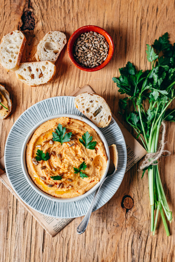 Astuces pour manger des graines au quotidien. Un bol de houmous crémeux, garni de paprika, d’un filet d’huile d’olive et de feuilles de persil frais, est présenté sur une assiette en céramique. Des tranches de pain croustillant entourent le plat, accompagnées d’un petit bol de graines de tournesol et d’un bouquet de persil noué avec une ficelle rustique, le tout disposé sur une table en bois.