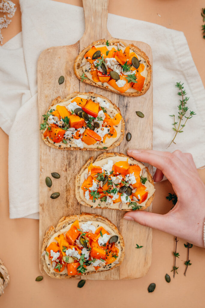 Astuces pour manger des graines au quotidien. Tartines gourmandes à la courge rôtie, fromage frais et graines sur une planche en bois.

