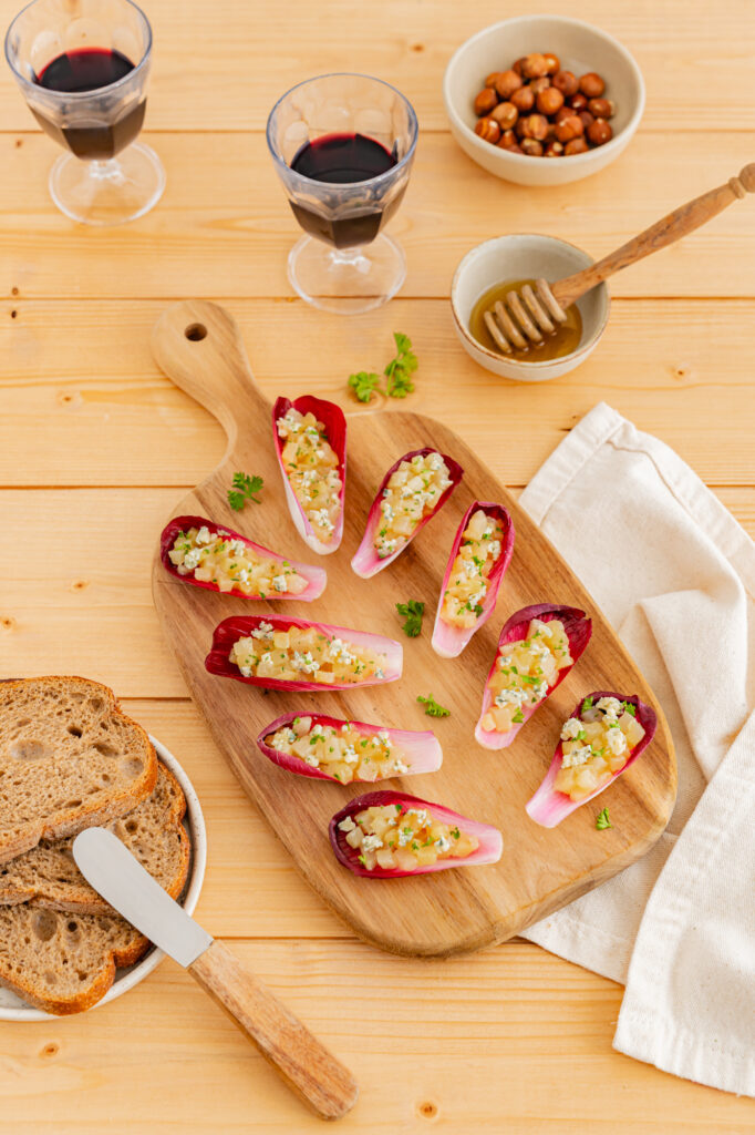 Amuse-bouches endives, poire bleu et miel. Recette à base de feuilles de chicorée garnies de fromage et de noix, disposés sur une planche en bois avec du pain et du miel.