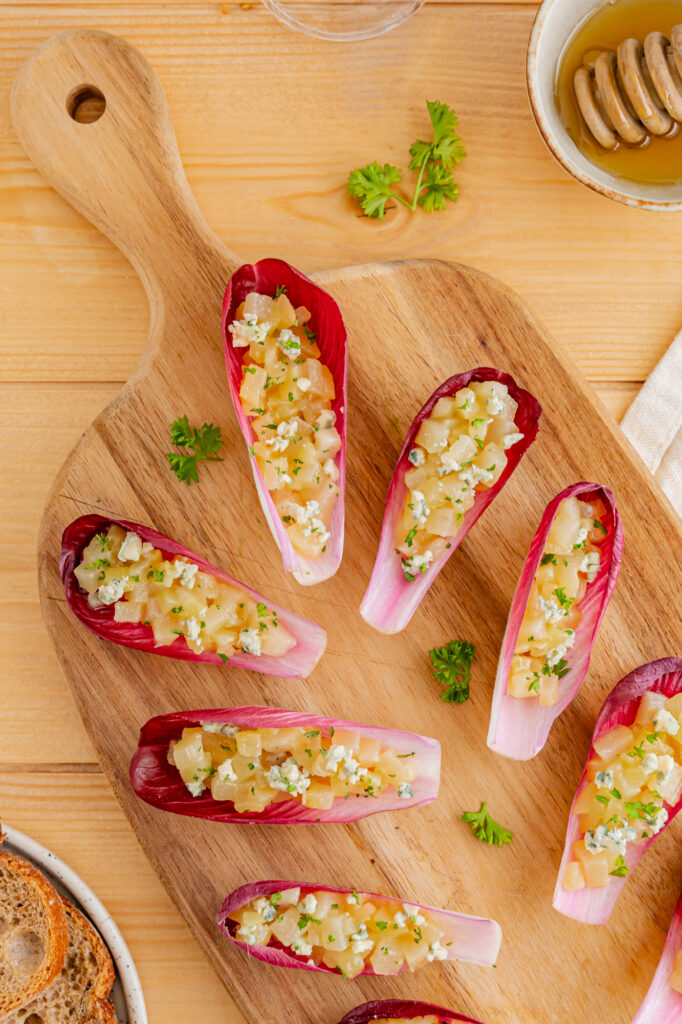 Amuse-bouches endives, poire bleu et miel. Recette à base de feuilles de chicorée garnies de fromage et de noix, disposés sur une planche en bois avec du pain et du miel.