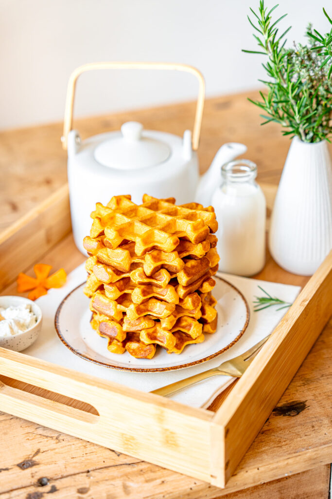Plateau en bois avec une pile de gaufres aux patates douces servies sur une assiette, accompagnées d’une théière blanche, d’une bouteille de lait et d’un brin de romarin.