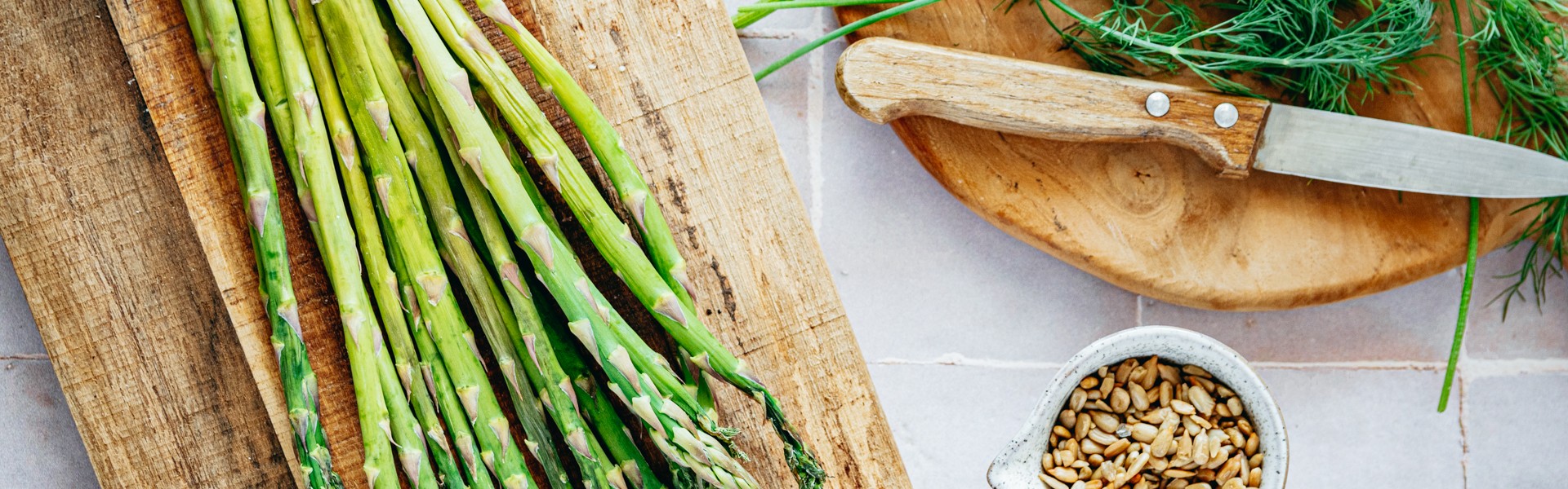 Les fruits et légumes du printemps. Asperges disposées sur une planche et un fond carrelage, accompagnée d'un couteau en bois, d'herbe aromatique et de pignons de pin. 
