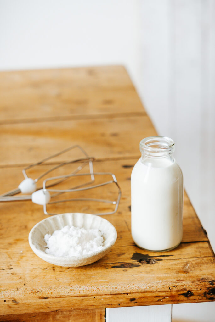 Astuces pour réussir et conserver sa chantilly. Ingrédients pour préparer une chantilly maison – crème fraîche, sucre glace et fouets de batteur sur une table en bois.